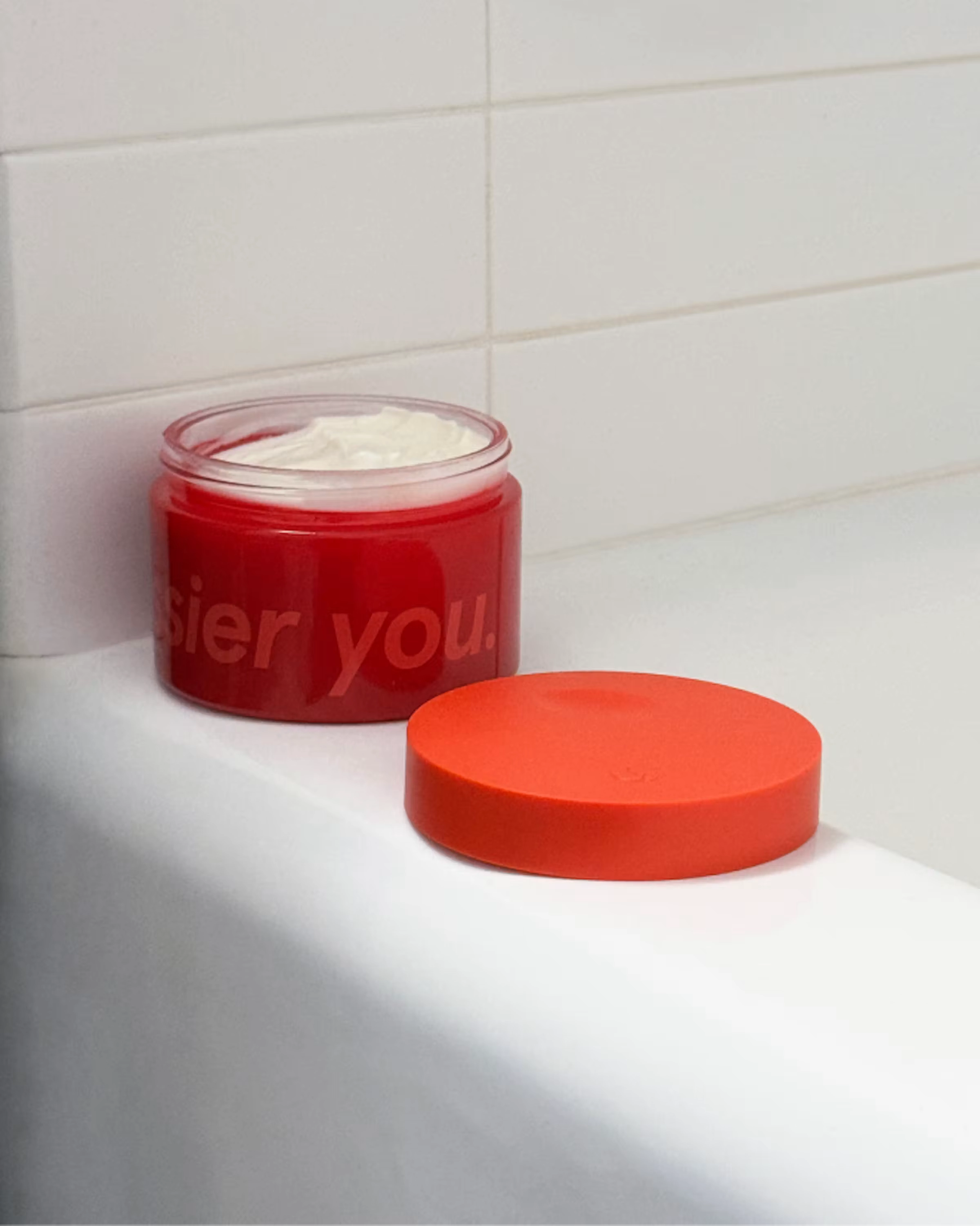 Red body cream jar with its lid beside it on a white ledge