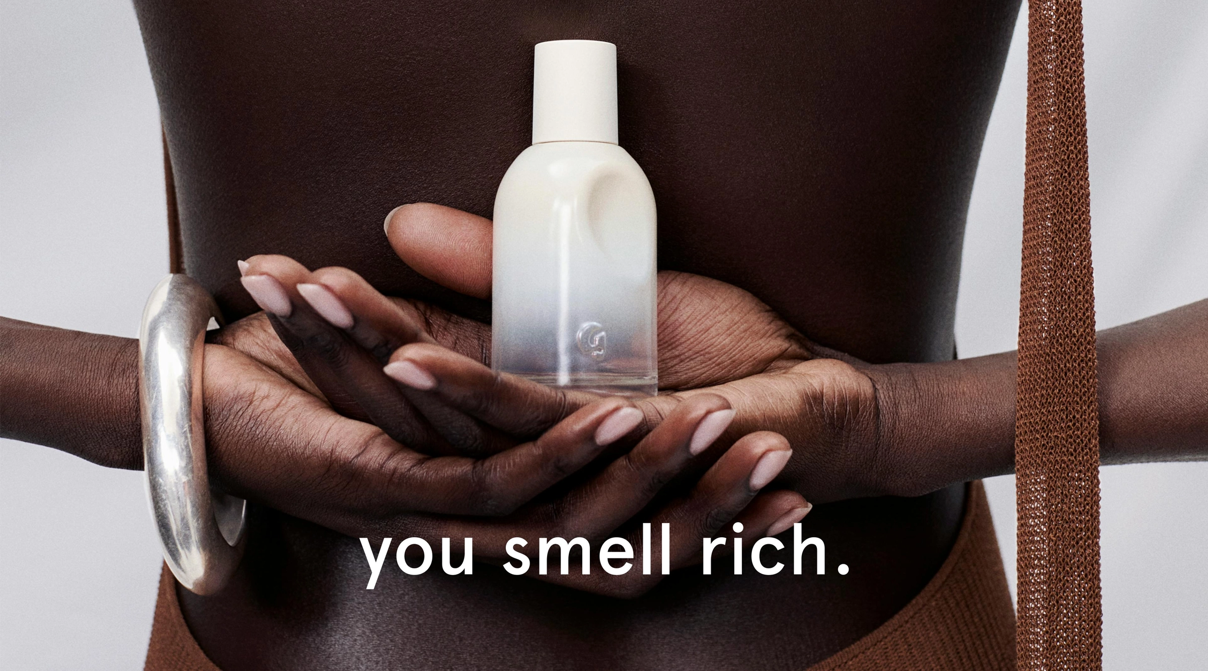Campaign photo from the Glossier home page: hands holding a white fragrance bottle against a dark torso, with the words you smell rich on the image